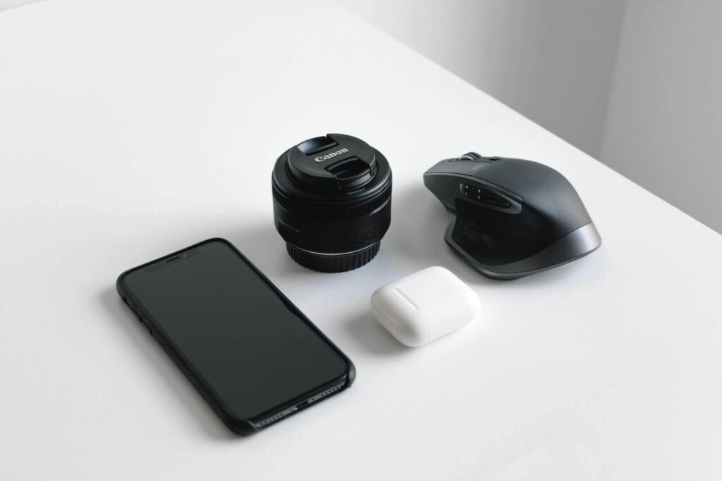 Телефон, наушники, мышка и объектив от фотоаппарата посреди белого стола