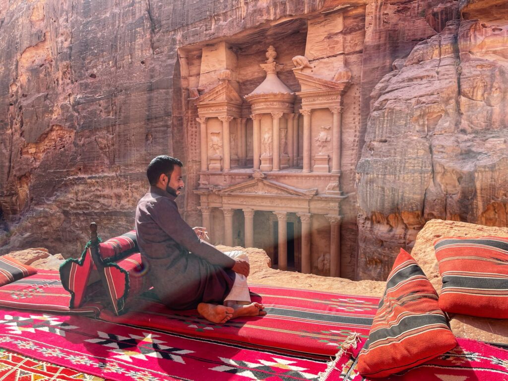 Человек смотрит на храмы Петры, сидя на красном ковре и опираясь на красное изголовье