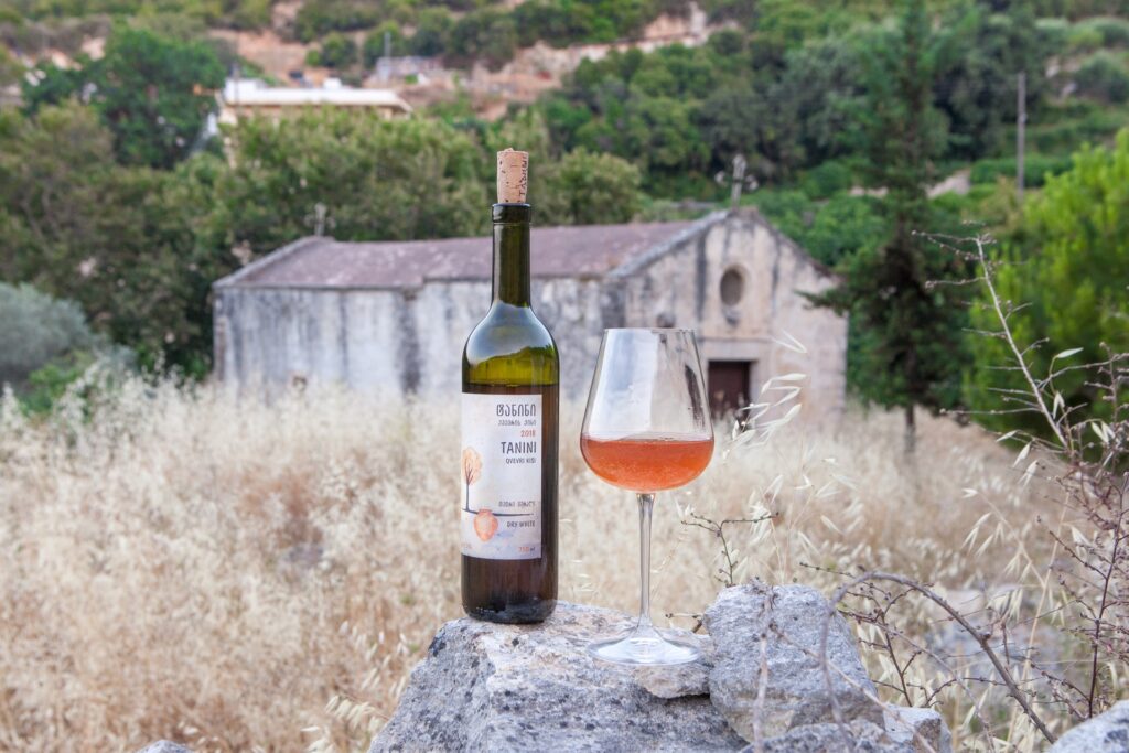 Бутылка и бокал с оранжевой жидкостью на фоне сельского дома